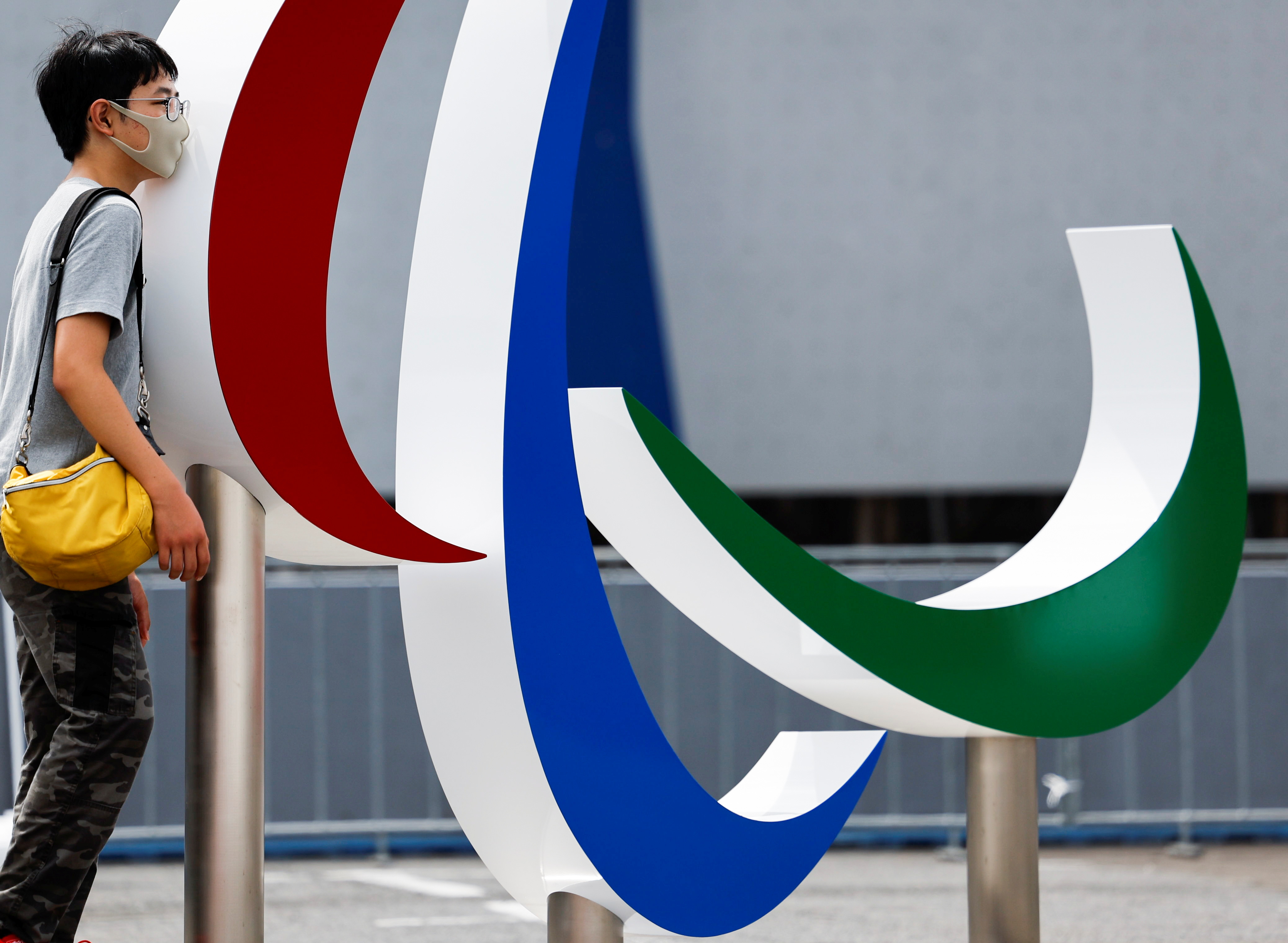 A visitor wearing a protective face mask poses for a photograph next to the monument of Paralympic symbol 'The three Agitos' in Tokyo