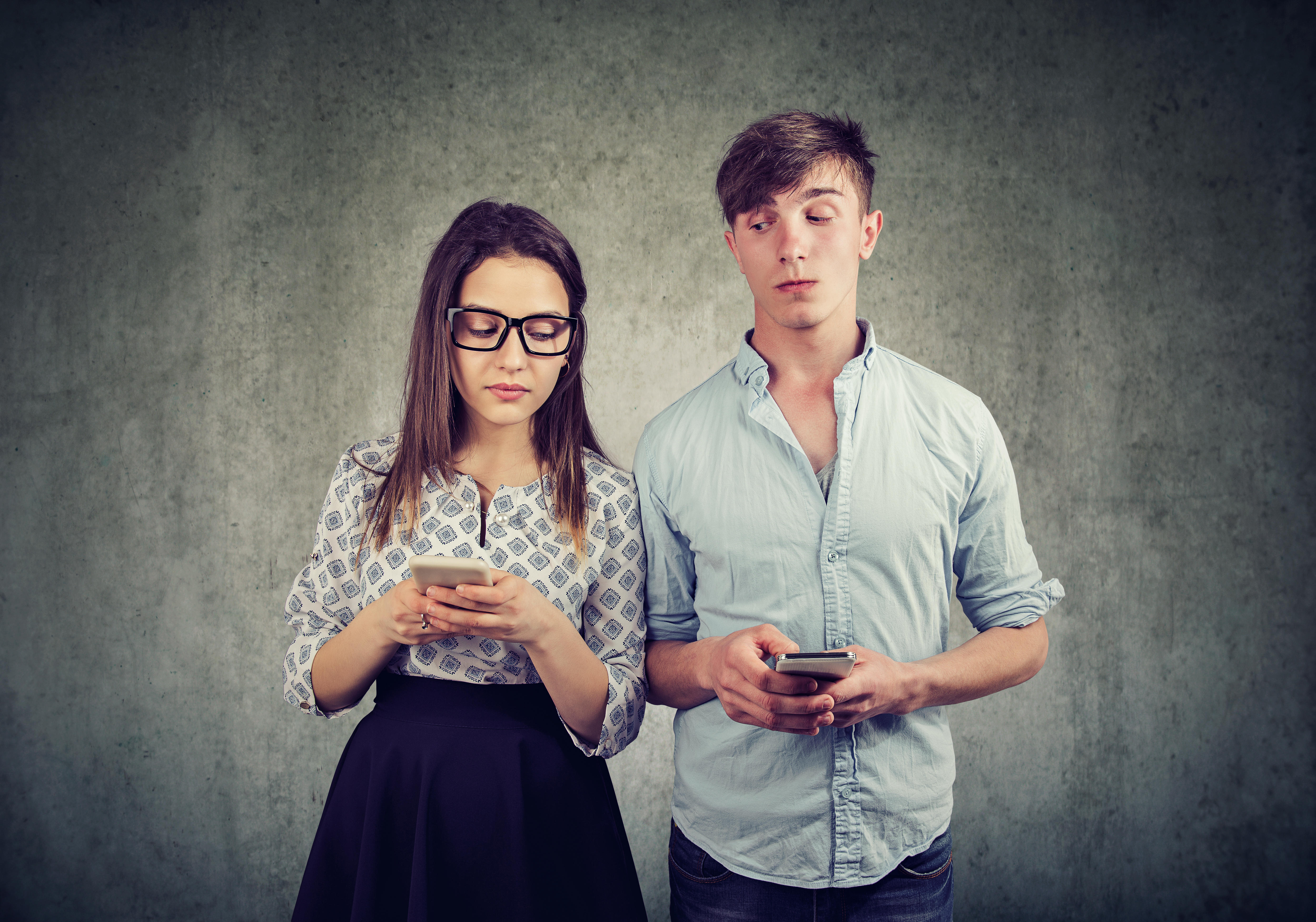 Young man and woman looking and peeking in smartphones of each other in disbelief